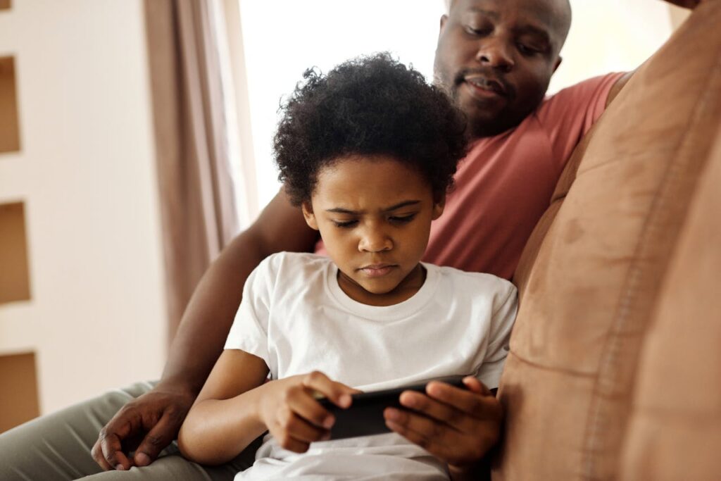 A father and son enjoy leisure time indoors while focusing on a smartphone.