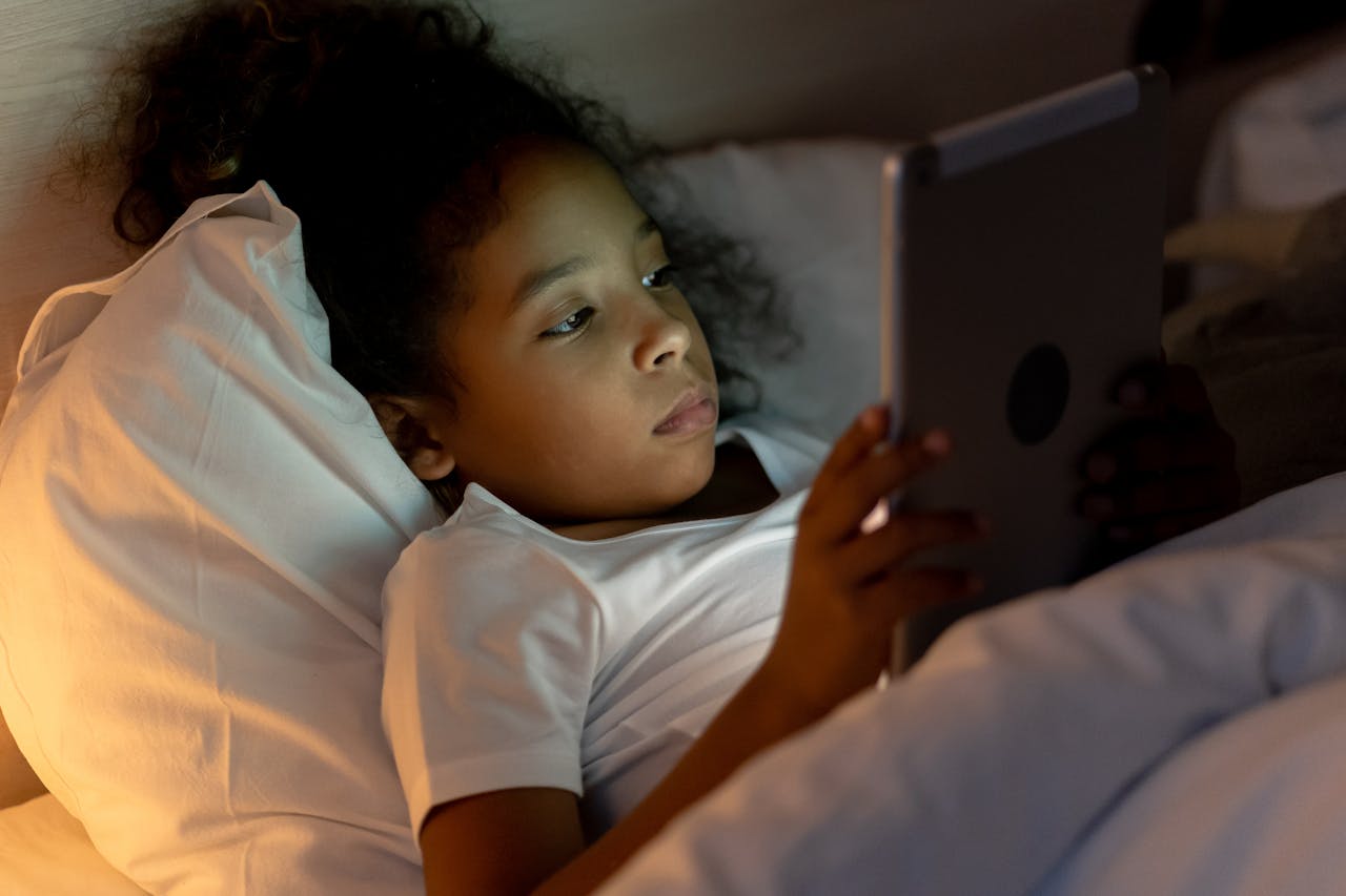 A young girl lying in bed, engrossed in a tablet screen at night, enjoying her bedtime routine.